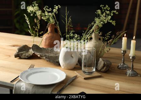 Set aus sauberem Geschirr, Vasen mit schönen Blumen, Steinen und brennenden Kerzen auf Holztisch im stilvollen Esszimmer Stockfoto