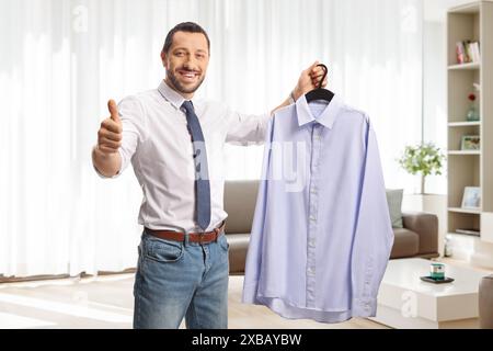 Ein Geschäftsmann hält ein Hemd an einem Kleiderbügel zu Hause im Wohnzimmer und bewegt die Daumen hoch Stockfoto