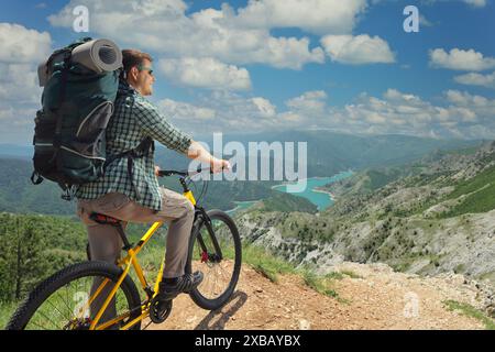 Ein Mann mit einem Fahrrad, der am Rande einer Bergklippe steht und auf einen See blickt Stockfoto