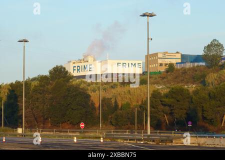 Barcelona, Spanien - 15. Mai 2023: Rauch tritt aus der FRIME-Fabrik hinter einer Baumlinie auf, während die Sonne untergeht, von einer leeren Autobahn aus gesehen. Stockfoto