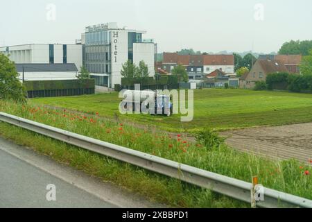 Gent, Belgien - 22. Mai 2023: Ein klassischer Traktor pflügt ein grünes Feld vor dem Hintergrund eines mehrstöckigen Hotelgebäudes und roter Mohnblumen Stockfoto