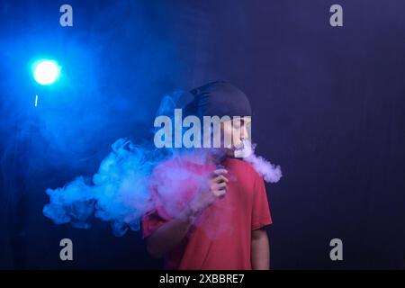 Porträt eines ernsten Reifen asiatischen Mannes in rotem T-Shirt, der E-Zigarettenrauch auf schwarzem Hintergrund ausatmet. Gesundheitsgefährdungskonzept. Stockfoto