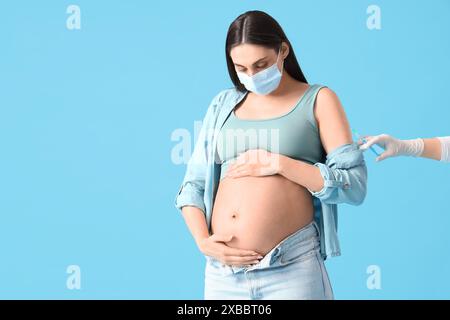 Junge schwangere Frau in medizinischer Maske, die Impfstoff auf blauem Hintergrund erhält Stockfoto