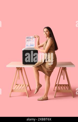 Junge Frau, die Dokumente mit dem Aktenvernichter auf dem Tisch vor rosa Hintergrund zerstört Stockfoto