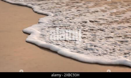 Eine entgegenkommende Welle in Bewegung. Genießen Sie den beruhigenden Anblick der ruhigen Strandwellen, die sich bei Sonnenuntergang sanft auf die Sandküste stürzen Stockfoto