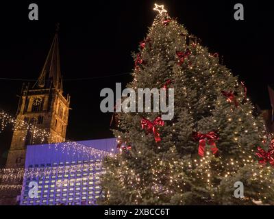 In der Mitte ein großer, festlich geschmückter Weihnachtsbaum mit Lichtern, roten Bögen und einem Stern an der Spitze, neben einer beleuchteten Kirche bei Nacht, dem Ahaus Stockfoto