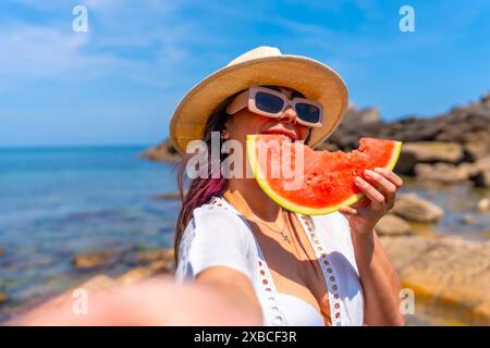 Eine Frau isst eine Wassermelone am Strand. Die Wassermelone wird in zwei Hälften geschnitten und sie hält sie in der Hand. Der Strand ist wunderschön und entspannend Stockfoto