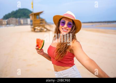 Eine Frau lächelt und hält eine Wassermelonenscheibe. Sie trägt einen Strohhut und eine Sonnenbrille Stockfoto