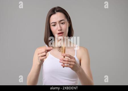 Eine gestresste Frau, die ihr verlorenes Haar von der Bürste auf grauem Hintergrund nimmt. Alopezie-Problem Stockfoto