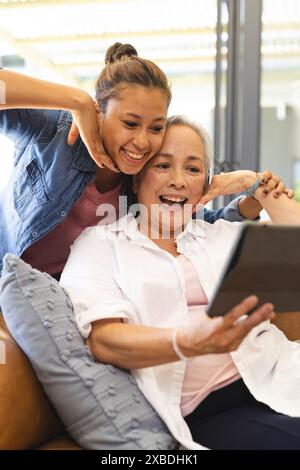 Asiatische Großmutter und birassische Enkelin mit Selfie-Tablette. Sie sitzen auf einer gemütlichen Couch in einem hellen, modernen Wohnzimmer mit großem Gewinn Stockfoto