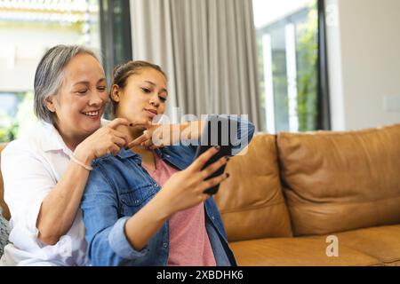 Asiatische Großmutter und birassische Enkelin machen Selfie auf einem Smartphone. Sie sitzen auf einer braunen Ledercouch in einem hellen, modernen liv Stockfoto