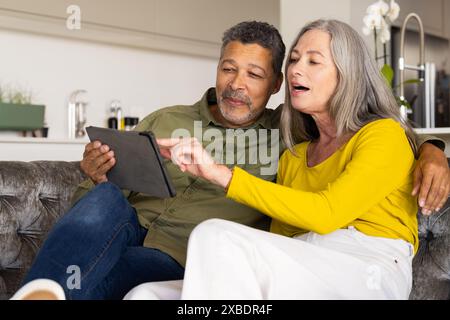 Ein geschlechtsreiches Ehepaar sitzt auf der Couch, interagiert mit einem Tablet, zu Hause. Sie sind in einem modernen Wohnzimmer mit Küche im Hintergrund, shari Stockfoto