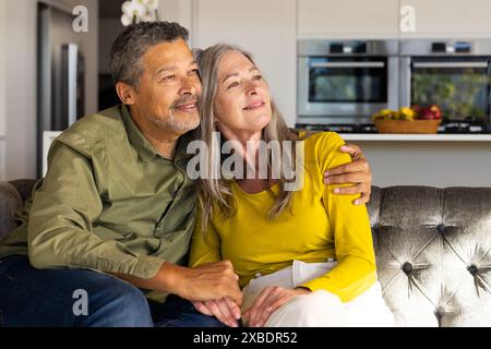 Ein zweirassiges reifes Paar sitzt auf der Couch, lächelt und sieht zufrieden aus, zu Hause. Sie sind in einer modernen Küche mit Edelstahlgeräten und einem Stockfoto