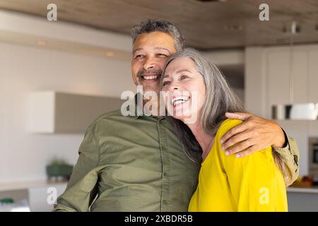 Ein birassischer reifer Mann und eine kaukasische reife Frau lächeln und umarmen zu Hause. Steht in einer modernen Küche mit weißen Schränken und Edelstahl Stockfoto