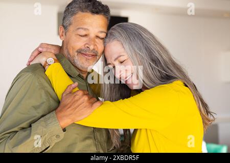 Zweirassiges reifes Ehepaar, das zu Hause, zu Hause, freundlich lächelt und umschließt. Helles und gemütliches Wohnzimmer mit moderner Einrichtung, hellem Hintergrund und Co Stockfoto