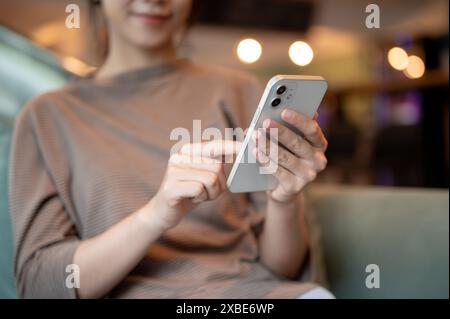 Ein zugeschnittenes Bild einer positiven jungen asiatischen Frau, die ihr Smartphone benutzt, während sie auf einem Sofa in einer Hotellounge oder einem Café sitzt. Personen und Wireless T Stockfoto