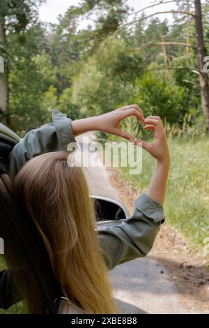 Frau, die im Auto fährt, die Hand aus dem Fenster hält und ein Herzsymbol mit ihren Händen zeigt. Love and Adventure Love Love and Adventure Local Travel Freiheit und Verbindung mit der Natur. Roadtrip mit nachhaltigem Lebensstil am Wochenende zum Entspannen und Ausruhen Stockfoto