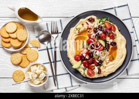 Griechischer Hummus mit frischen Gurken, Oliven, Tomaten, Fetakäse, roten Zwiebeln und Thymian auf einem Teller auf weißem Holztisch mit Crackern, Löffel und Stockfoto