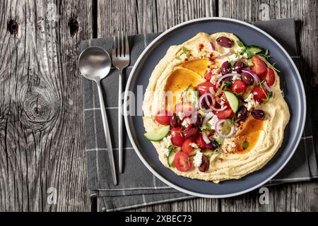 Griechischer Hummus mit frischen Gurken, Oliven, Tomaten, Fetakäse, roten Zwiebeln und Thymian auf Teller auf rustikalem Holztisch mit Löffel und Gabel, Hor Stockfoto