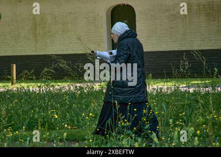 Brügge, Brüssel; Juni 2024; eine alte Nonne spaziert durch den Klostergarten. Mit gewalztem Material beladen. Gekleidet mit schwarz-weißem Verschluss Stockfoto