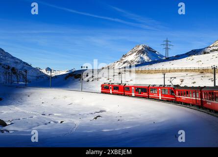 Berina, Schweiz - Januar 06. 2023: Die rote Rhätische Bahn fährt entlang des gefrorenen Bianco-Sees in einem dramatischen Sonnenloch, mit den Alpen summ Stockfoto