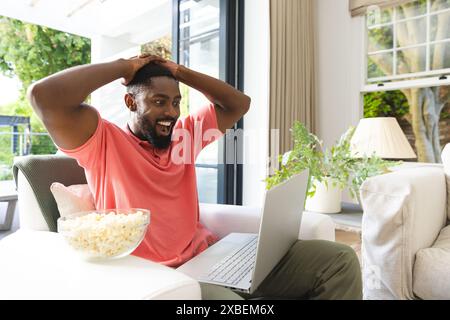 Aufgeregt afroamerikanischer Mann, der etwas auf seinem Laptop sieht, zu Hause. Helles Wohnzimmer mit großen Fenstern und Popcorn-Schale auf dem Tisch, unverändert Stockfoto