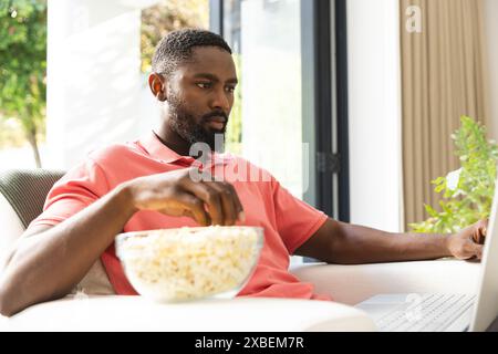 Junger afroamerikanischer Mann, der sich zu Hause entspannt, Popcorn isst, während er Laptop benutzt, zu Hause. Helles, modernes Wohnzimmer mit großen Fenstern und Grünflächen Stockfoto