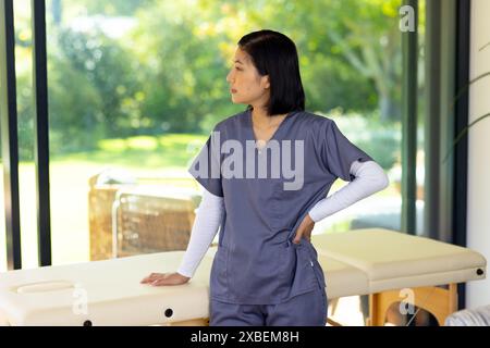 Asiatischer Physiotherapeut steht im Zimmer mit Massagetisch und trägt medizinische Peelings. Die großen Fenster bieten einen üppigen, grünen Garten im Freien, der eine Atmosphäre schafft Stockfoto