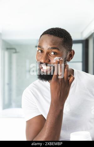 Junger afroamerikanischer Mann, der Gesichtscreme aufträgt, warm lächelt, zu Hause. Helles, modernes Badezimmer mit Glasdusche im Hintergrund, weißen Fliesen und s Stockfoto