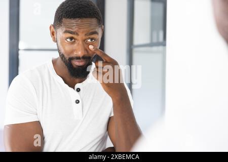 Junger afroamerikanischer Mann mit Bart, der sein Gesicht berührt, während er in einen Spiegel blickt, zu Hause Stockfoto