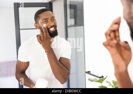 Junger Mann aus Afroamerikaner, der Gesichtscreme aufträgt, während er sein Spiegelbild anlächelt, zu Hause. Modernes Bad mit Glastüren und Handtuch im Hintergras Stockfoto