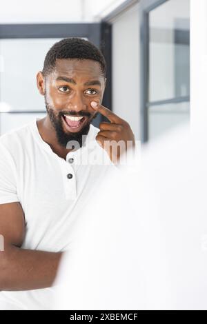Junger Mann aus Afroamerikaner mit Bart, der auf sein Gesicht zeigt und lächelt, zu Hause. Helles, modernes Innenleben mit großen Fenstern im Hintergrund, kreativ Stockfoto