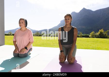 Asiatische Großmutter und birassische Enkelin üben gemeinsam Yoga auf Matten. Sie sind im Freien mit malerischem Blick auf die Berge und üppig grünem Rasen hinten Stockfoto