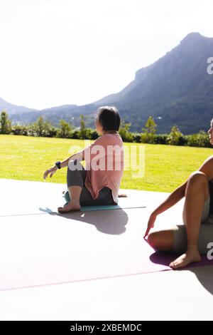 Asiatische Großmutter und birassische Enkelin üben gemeinsam Yoga im Freien. Sonniger Tag mit Bergen im Hintergrund, beide genießen die frische Luft und Stockfoto