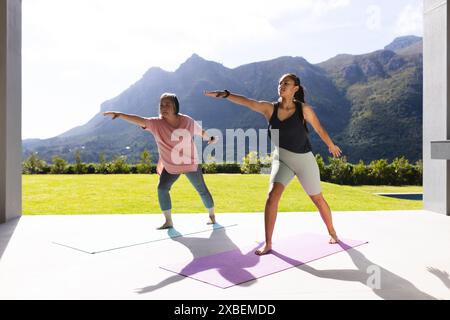 Asiatische Großmutter und birassische Enkelin üben gemeinsam Yoga auf Matten. Sie sind draußen mit Bergen und Grün im Hintergrund, genießen s Stockfoto