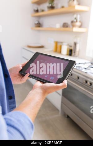 Junger kaukasischer Mann, der ein Tablet hält und eine Smart Home App-Schnittstelle anzeigt. Moderne Küche mit Regalen und Geräten in weißem Hintergrund, zeigt Stockfoto