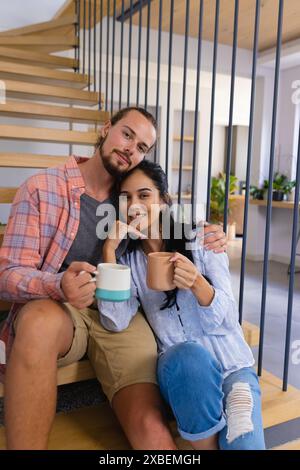 Junges, vielfältiges Paar, das auf der Treppe sitzt und Kaffeetassen hält. Moderne Inneneinrichtung mit Holzakzenten und offenem Design im Hintergrund, unverändert Stockfoto