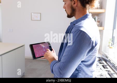 Junger kaukasischer Mann mit Smart Home App auf Tablet in einer modernen Küche. Helles Sonnenlicht, das durch große Fenster strömt, beleuchtet elegante, minimalistisch Stockfoto