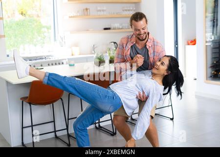 Junge, vielfältige Paare tanzen spielerisch in einer modernen Küche. Helles Tageslicht erfüllt den Raum und unterstreicht die eleganten Arbeitsflächen und die stilvolle Einrichtung, unalt Stockfoto