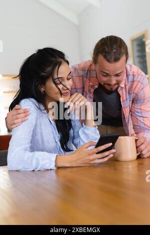 Eine junge Frau schaut auf ihr Smartphone, während ein junger Kaukasier neben ihr steht. Sie sind in einem modernen Haus mit einem Holztisch und einem Cof Stockfoto