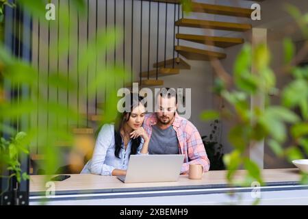 Ein vielseitiges Paar, das zusammen sitzt und auf den Laptop-Bildschirm blickt. In einem modernen Haus mit einer stilvollen Treppe und Grün, die draußen sichtbar ist, schaffen eine ruhige Atmosphäre Stockfoto