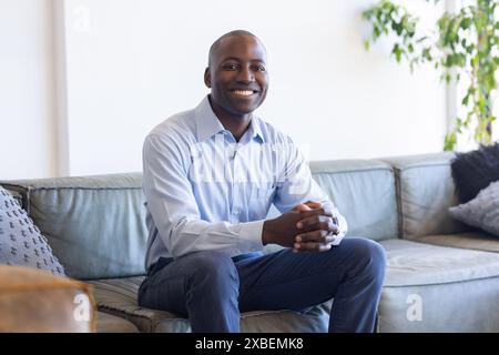 Porträt eines jungen afroamerikanischen Mannes, der auf der Couch sitzt und selbstbewusst lächelt. Helles Wohnzimmer mit moderner Einrichtung und Topfpflanze im Hintergrund, Stockfoto