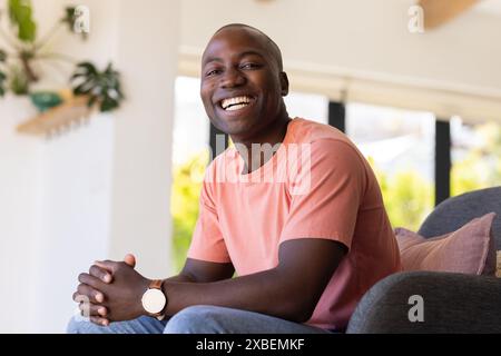 Porträt eines jungen afroamerikanischen Mannes, der lächelnd auf der Couch in einem hellen Wohnzimmer sitzt. Große Fenster und grüne Pflanzen im Hintergrund erzeugen ein Stockfoto