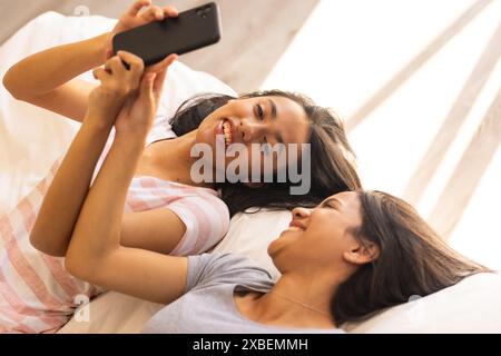 Zwei junge birassische Schwestern liegen auf einem Bett, lächeln und teilen sich einen Moment mit einem Telefon, zu Hause Stockfoto