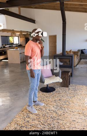 Junger afroamerikanischer Mann mit Virtual-Reality-Headset im modernen Wohnzimmer. Offener Raum mit Holzdecke, gemütlichen Möbeln und großem Wind Stockfoto