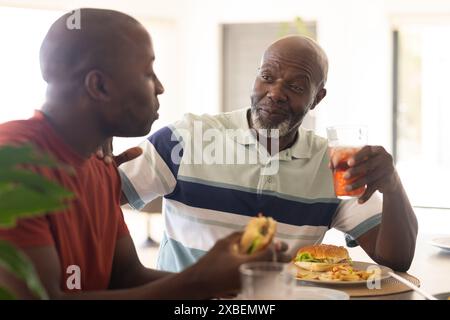 Afroamerikanischer Vater und Sohn genießen zu Hause zusammen ein Essen. Heller Speisesaal mit Tageslicht, das durch große Fenster strömt, sorgt für eine warme Atmosphäre Stockfoto