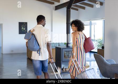 Junges vielseitiges Paar, im Ferienhaus mit Gepäck. Sie sind in einem modernen, gut beleuchteten Ferienhaus mit großen Fenstern und zeitgenössischen Möbeln, u Stockfoto