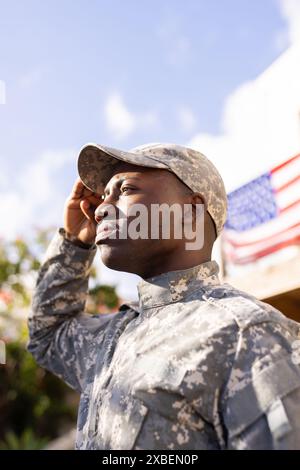 Junger afroamerikanischer Soldat in Militäruniform, der grüßt. Amerikanischer Flaggenhintergrund mit Outdoor-Grün, die patriotische Szene schafft, unverändert Stockfoto