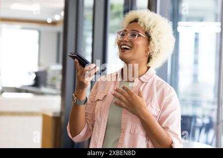Junge birassische Frau, die Smartphone hält und im Büro lächelt. Glastrennwände und moderne Einrichtung schaffen einen eleganten, unveränderten Hintergrund Stockfoto