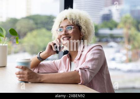 Junge birassische Frau mit lockigem Haar hält Kaffeetasse, spricht auf Smartphone im Geschäftsbüro Stockfoto